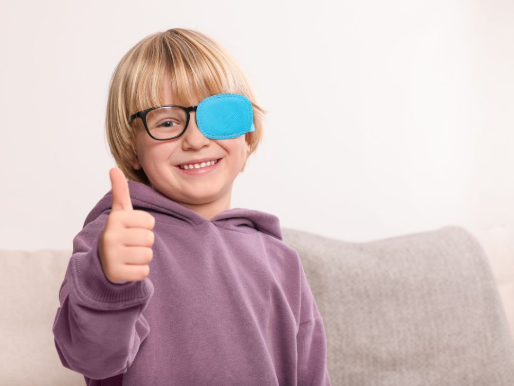 Criança com tapa-olho azul sorrindo e fazendo sinal de positivo, simbolizando o tratamento de condições relacionadas ao que causa estrabismo.